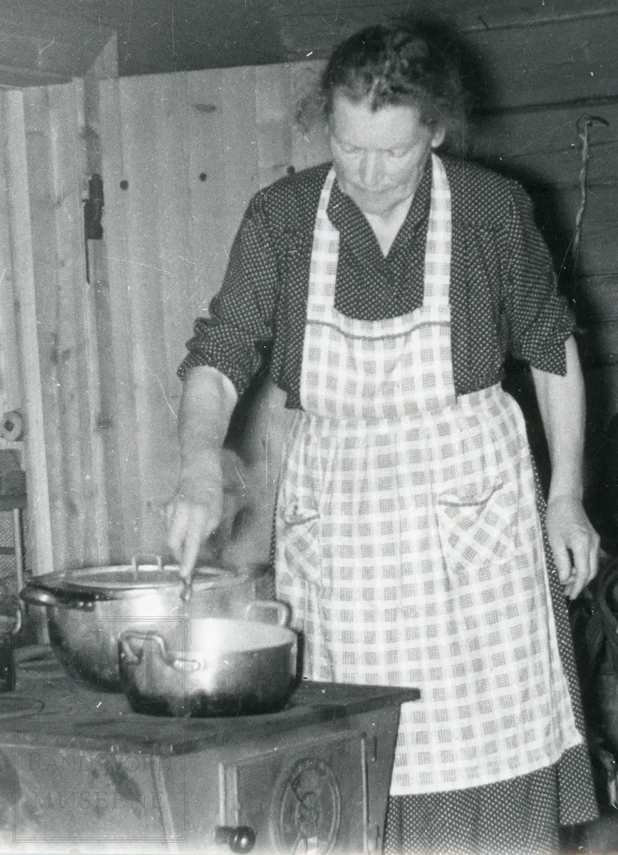 Kvinne med forkle står ved en ovn og rører i en kjele.