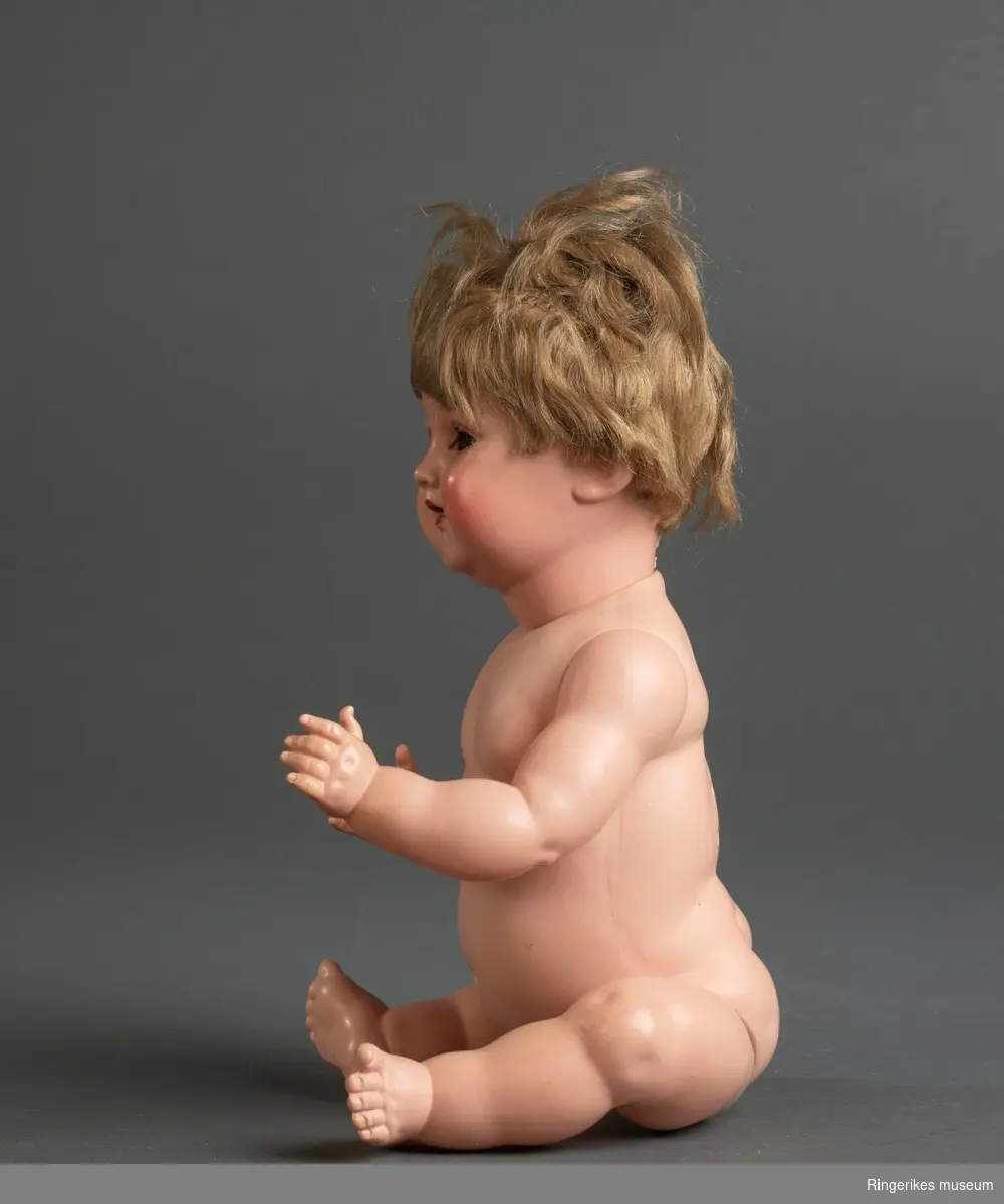 Dukke med babykropp, bøyde armer og ben, øyne som kan åpnes/lukkes og tykt hår. I kroppen lagt inn en boks som gir fra seg skrikelyd ved bevegelse.