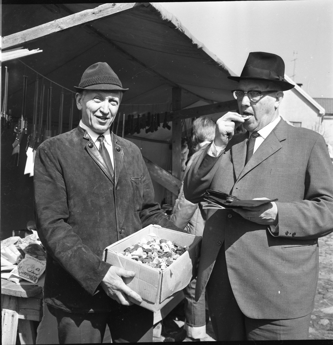 Höstmarknad i Gränna 1968. Gunnar Fransson - Gunnars-konfektyr - i hatt och mockajacka håller fram en kartong med karameller för marknadsföreståndaren (och polisen) Arvid Lindgren som låter sig smaka med plånboken uppslagen i näven. Det är möjligt att Lindgren, i hatt och kostym, går runt för att ta upp marknadsavgiften.
