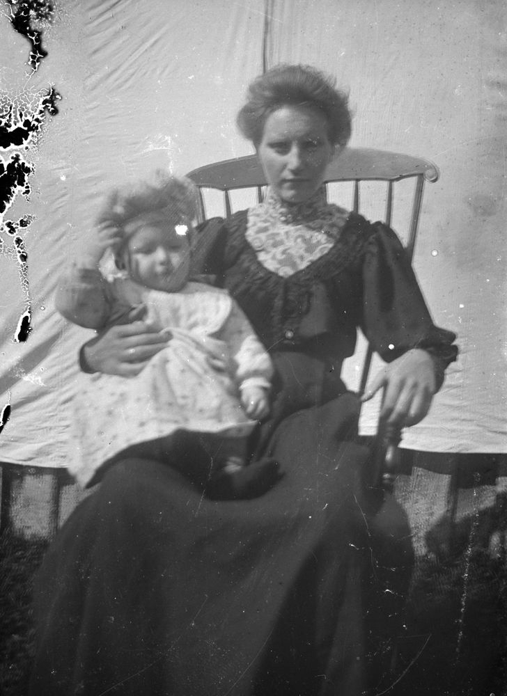 Leirfjord. Emma Johanne Aas Nicolaisdatter (f. 1883 - d. 1947) med lite barn på fanget, fotografert ute i en gyngestol foran et hvitt bakteppe. Finnes også på LEF.F.01324.