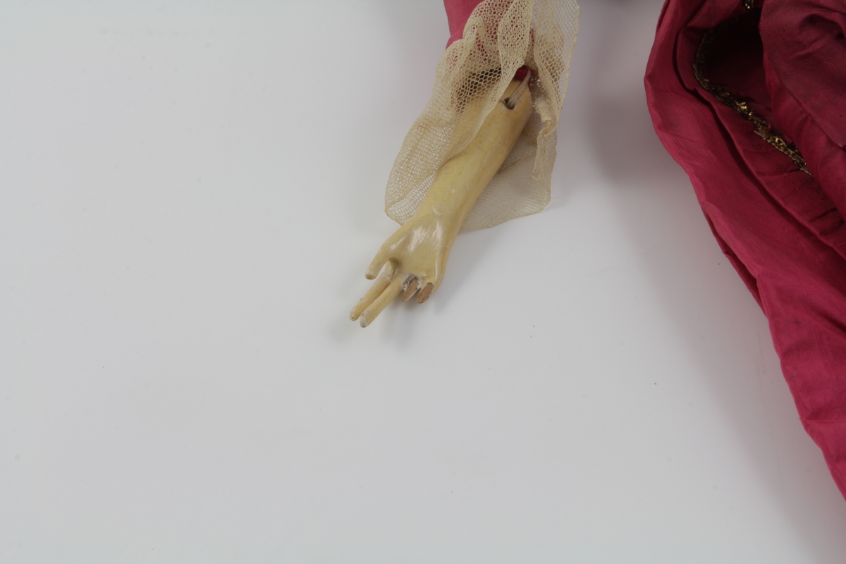 Dukke med kropp av tre. Hode og armer er dekket med et ytterlag, trolig gips, som er malt og glassert. Dukken har hår på hodet med en heklet kyse med rosa sløyfe over. Dukken er kledd i en rød ytterkjole i silke, ser hjemmelaget ut. Under denne er det en kjole av lin med broderte blomster. Under denne kjolen er en ny linkjole med en bord langs den nederste kanten med mørkeblå blomster og skrå røde striper. Innerst har dukken en enkel linkjole.