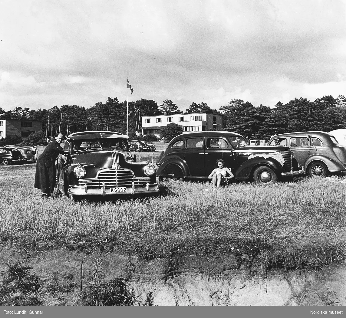 Parkerade bilar på en parkeringsplats vid en strand. En man står vid en Pontiac årsmodell 1946 och en kvinna sitter i en solstol vid en Chrysler årsmodell 1938.