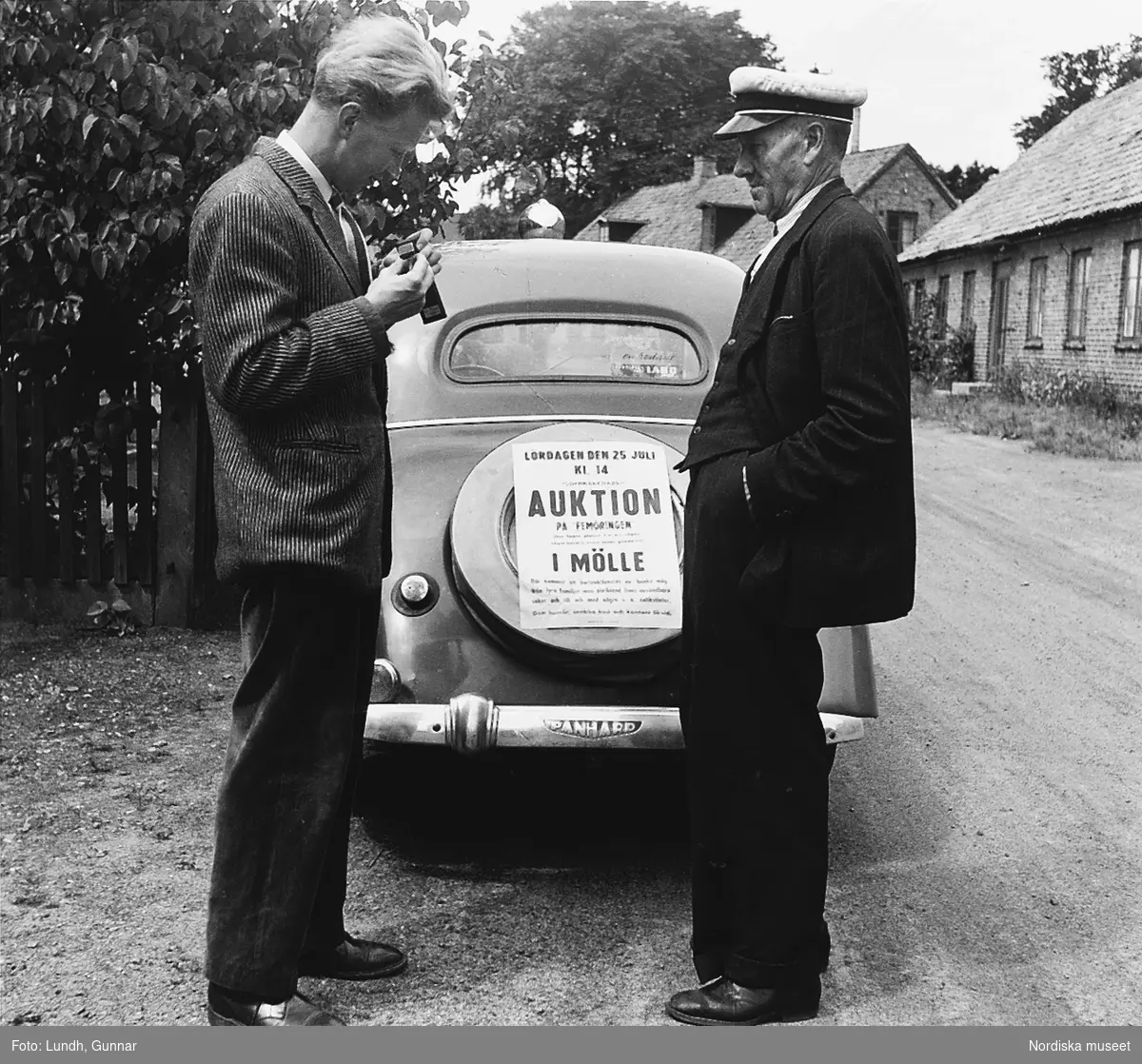 Två män står vid en bil i Mölle år 1953. På bilen sitter en affisch som gör reklam för "Auktion på Femöringen i Mölle".