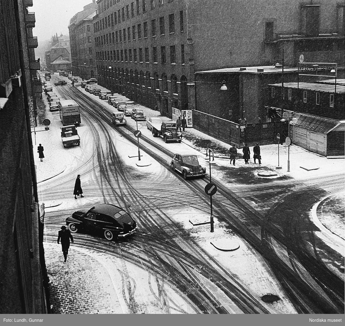 Utsikt från fotograf Gunnar Lundhs hem, Döbelnsgatan, Stockholm år 1955. Marken är snötäckt. Bilar och fotgängare på gatan.
