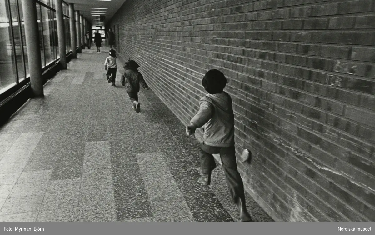 Barn springer efter varandra i lång korridor med tegelvägg på ena sidan och fönsterglas på den andra. Stengolv.