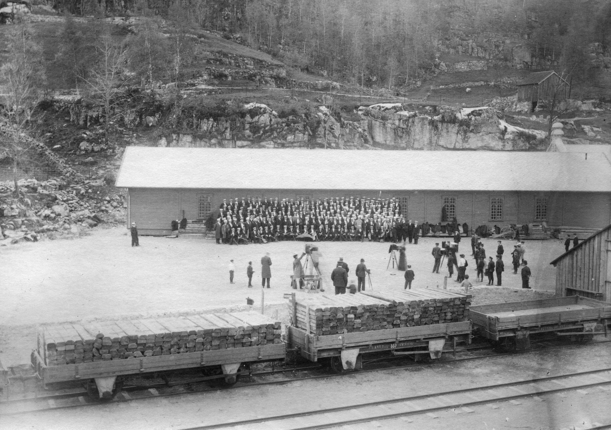 Flekkefjord 22.05.1904. Oversiktsbilde, kor og korps er oppstilt på Flekkefjord togstasjon under et sangerstevne for bildetaking. Tre kamera er oppstilt i front, to kvinnelige fotografer.