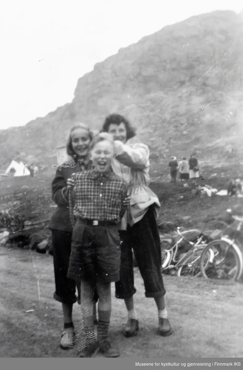 Nordvågen. St. Hans. Tre ungdommer på veien, i bakgrunnen hvite spisstelt, flere personer og noen sykler. 1953.