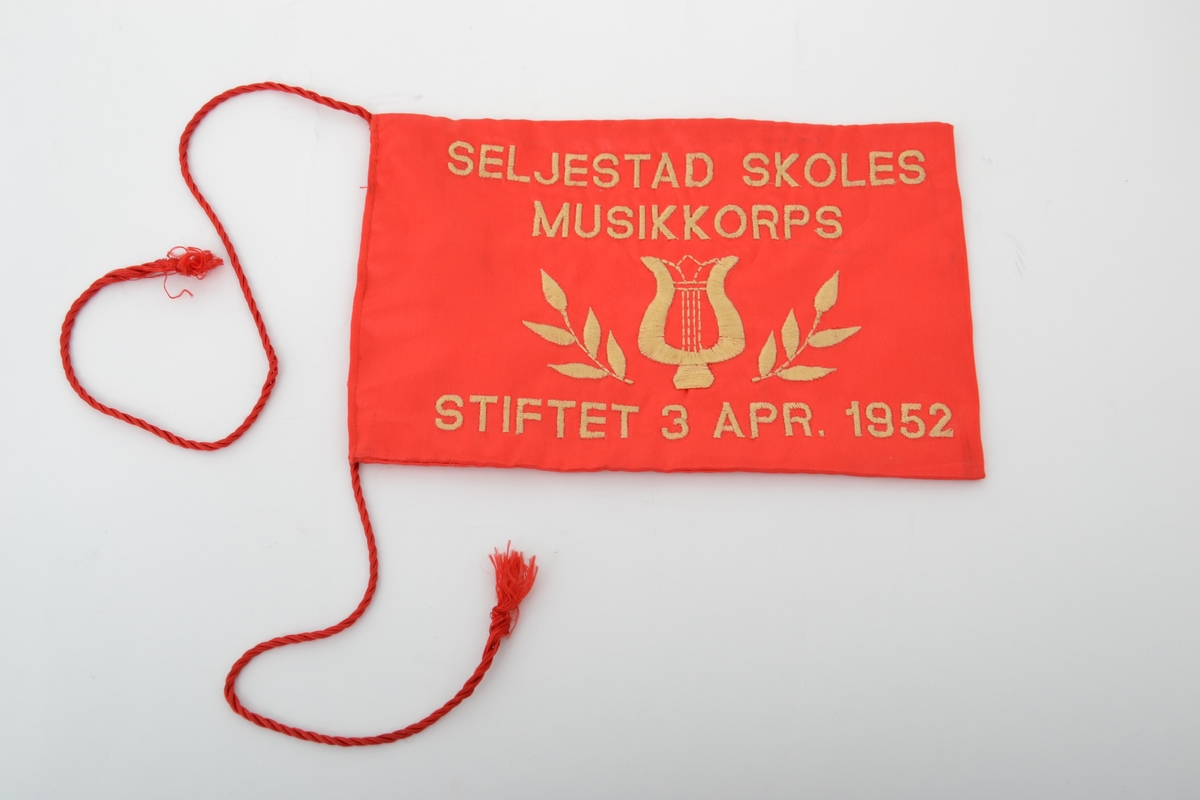 Bordfane fra Seljestad Skoles Musikorps i Harstad i rød silke brodert med gul tråd: en musikkharpe og bladmønster  med brodert tekst over