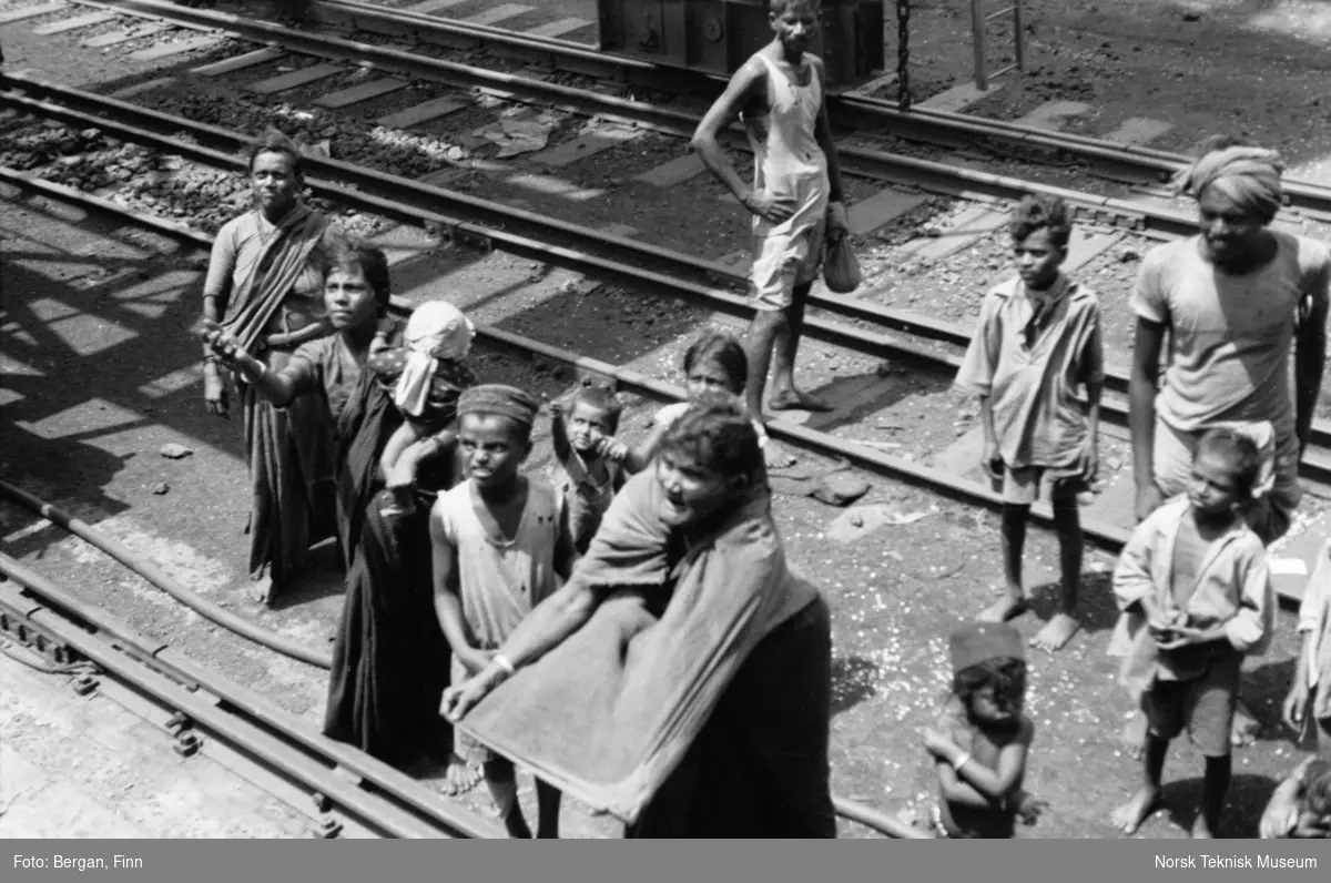 En gruppe voksne og barn samlet ved jernbanelinje, noen strekker hendene fram