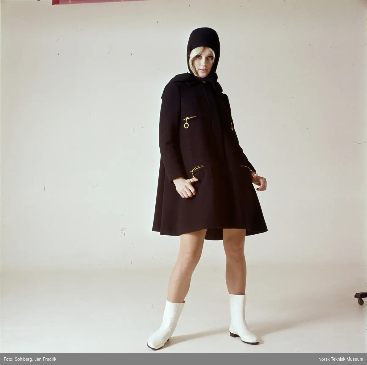 Tidlig norsk motefotografi. Kvinnelig modell poserer i brun kappe og lue.