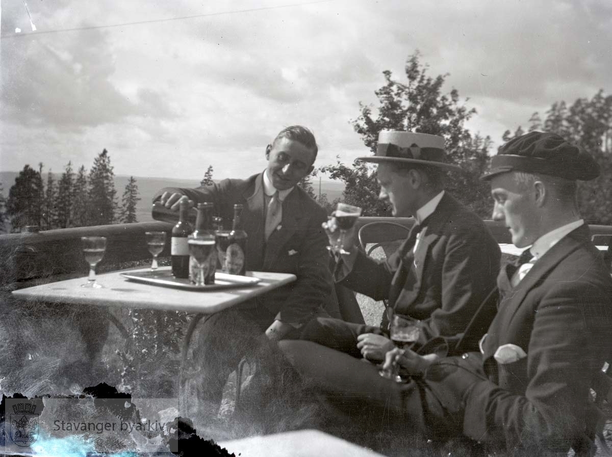 Tre menn drikker øl (?) på veranda.Flasker og glass.
