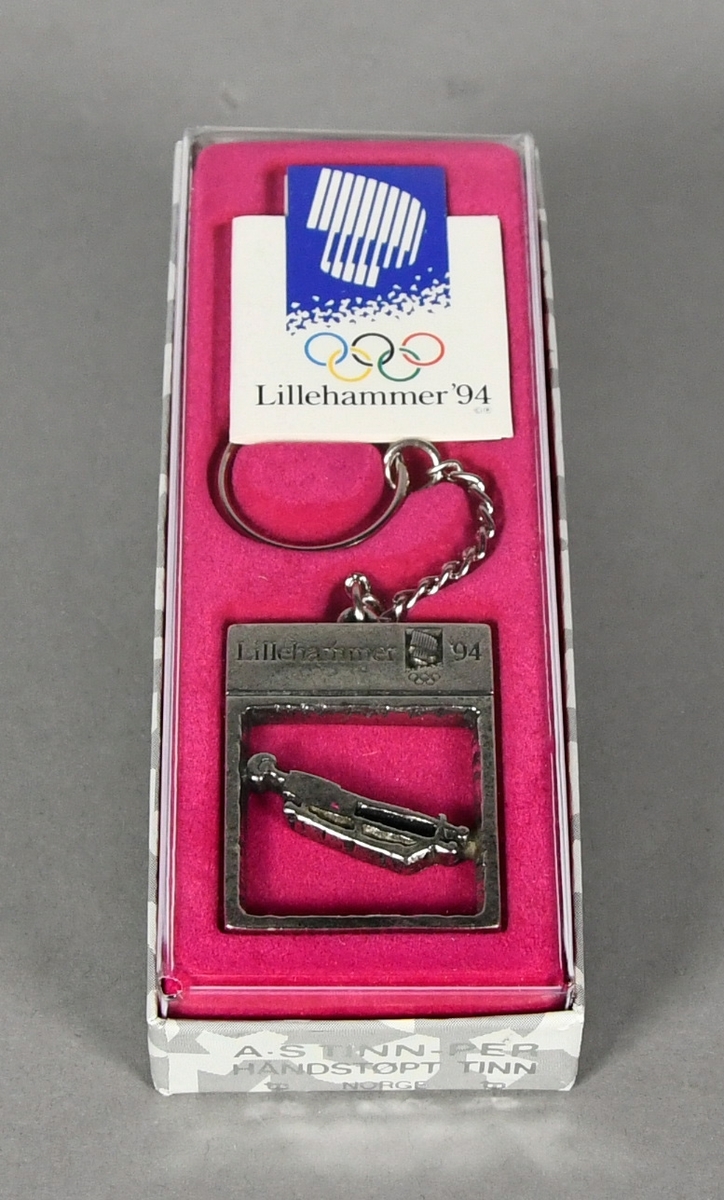 Nøkkelring med rektangulært anheng med piktogram for aking. Nøkkelringen ligger i original emballasje på rosa underlag.