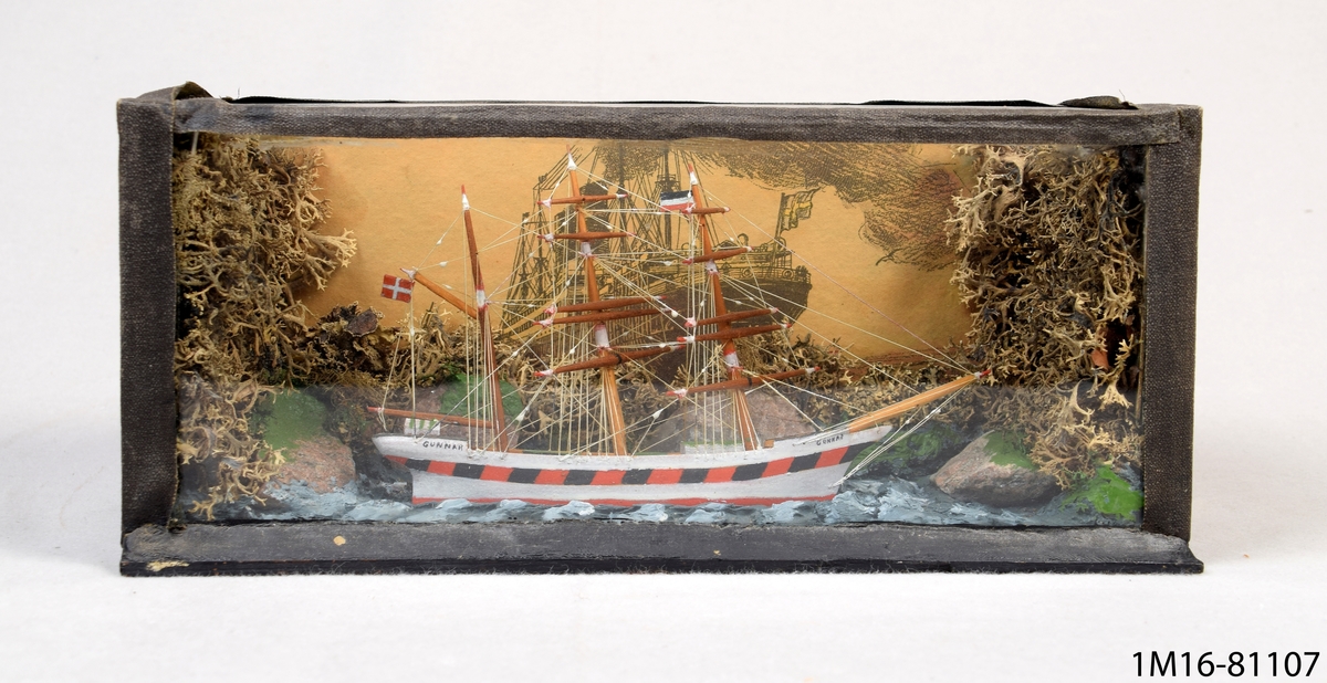 Liten båt, segelfartyg med tre master, i låda med glas framtill och upptill. 29½ x 10½, vid ändarna och baktill landskap av sten och mossa.