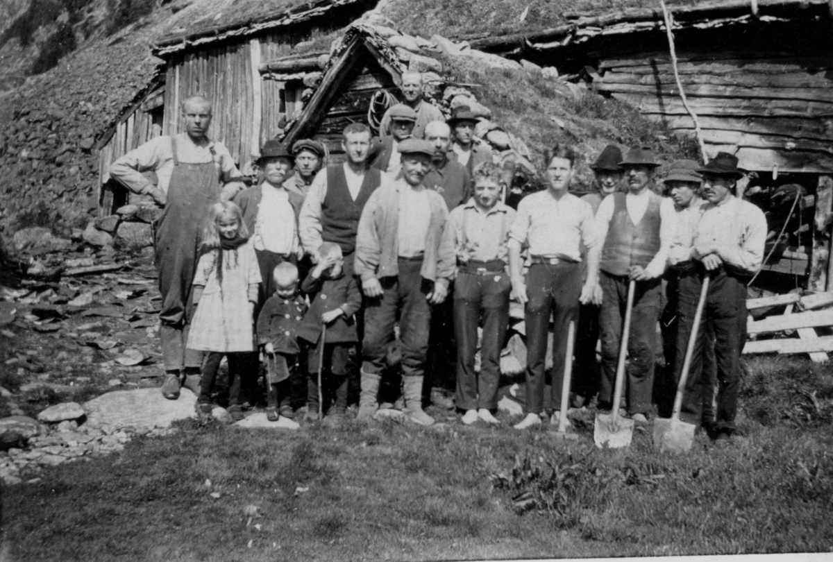 En gruppe menn og tre barn står foran et lite bruk i Ulladalen. Mennene er kledd i arbeidsklær og noen har spader.