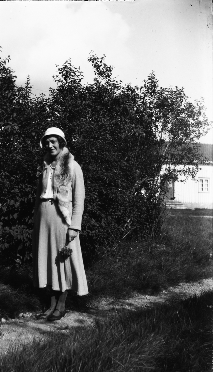 Ellen i Kalvhult står finklädd vid ett syrenbuskage med en kvist i handen. I bakgrunden syns ett bostadshus. Hon har en klockad hatt, pärlhalsband och rävstola.  (Se även bildnr EA0462)