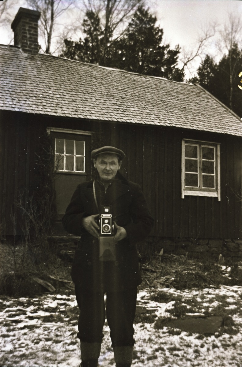Erik "Kloar-Erik" Andersson står med kameran framför sin stuga i skogen. Marken är lätt snötäckt.