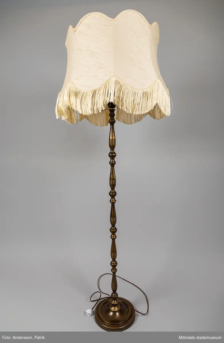 Golvlampa med skärm, tillverkad under 1970-talet. Lampfoten är profilerad och gjord av metall. Lampskärmen är i vit textil med fransar nedtill.