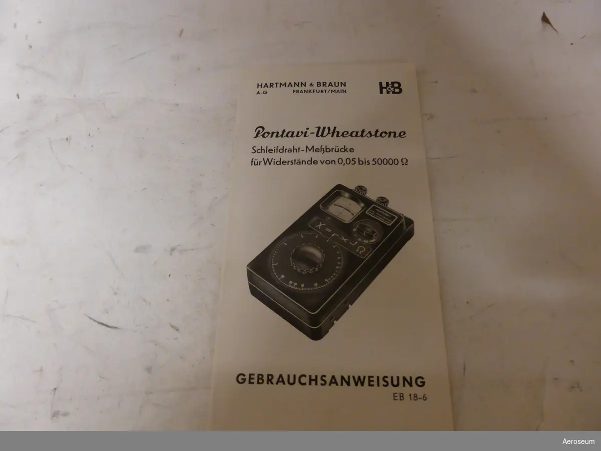 En Resistansmätbrygga, förvarad i en sliten blå pappersask. Runtom asken står det "ÖMTÅLIGT" på tejp. Tillverkad av Hartmann & Braun. På instrumentet finns en varningstext på tyska, och i asken finns även en bruksanvisning på tyska. I föremålets display står det: "H&B Pontavi Wheatstone".