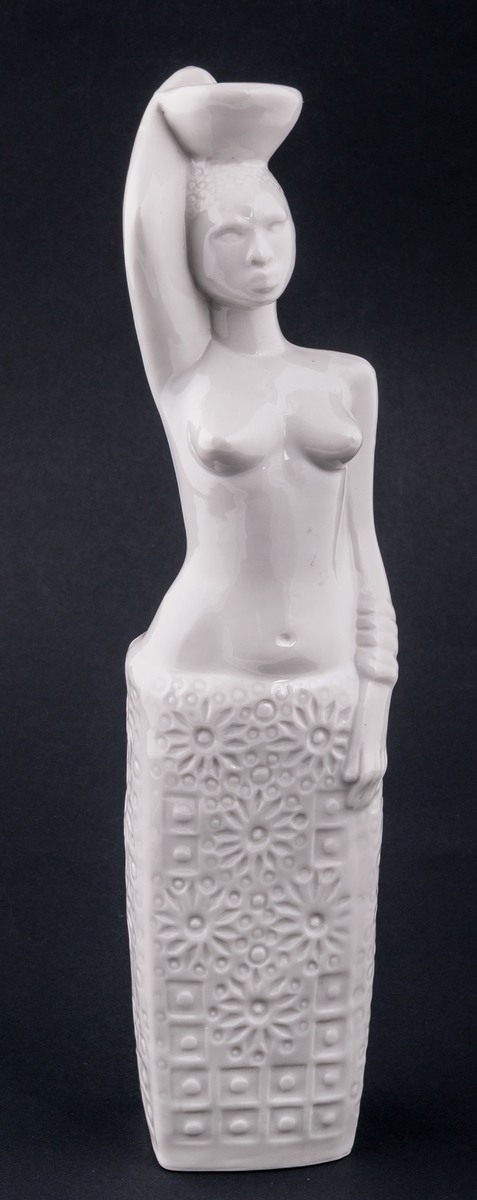 Figurin, föreställande en kvinna, bärandes kärl på huvudet, bar överkropp. Vit glasyr. Saknar stämplar och signatur. Okänd formgivare. Oklart om den är tillverkad vid Gefle Porslin eller Rörstrands porslinsfabrik.