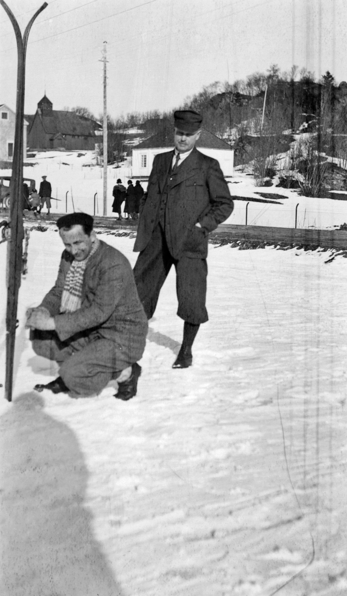 Vinterbilde med Trøndelag Folkemuseum i bakgrunnen. Stående ses Christian Anker Bachke.
"Mars 1934" påskrevet på baksiden.