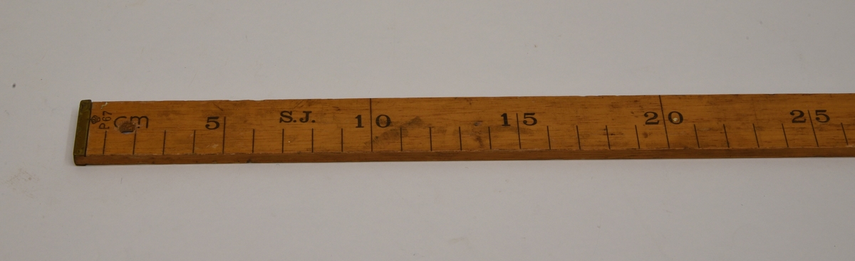 En trälinjal på 100 centimeter försedd med mässingsbeslag i båda ändar.

Linjalen är på framsidan märkt med "S.J." samt "P67" som också pryds med en krona. På baksidan är "Brastad" skrivet för hand med en tuschpenna, denna text är nött och lite svår att urskilja. På baksidan är det med en kulspetspenna också skrivet "90 x 35 x 55". I ena änden av linjalen finns  ett hål för att kunna fästa ett snöre vid den eller för att kunna hänga upp den på en spik på väggen, till exempel.