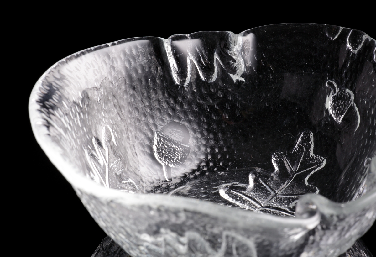 Asymmetriskt skål i ofärgat glas med reliefdekor av eklöv och ekollon.
Skålen är först gjuten till en "skiva" genom att man hällde ut glas över ett underlag med nämnda ekmönster. Sedan formades skålen i varmt tillstånd.