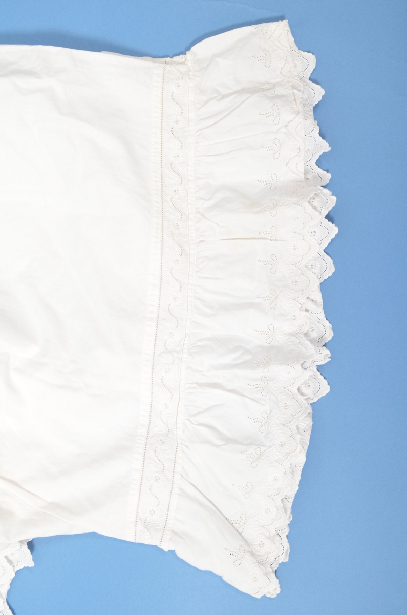 Underbukse i tynn kvit bomullslerrét. Dekor i sidene nederst på buksebeinet (strumpen) av engelsk broderi og tyll. Opning i kvar side, 32 cm lang.