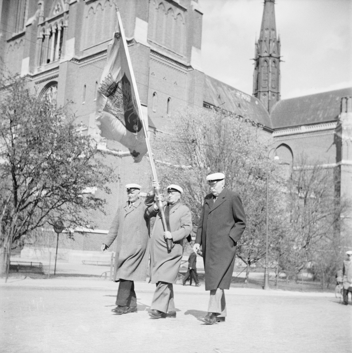 Tre män med studentmössor och fana gående vid Uppsala domkyrka, Östgöta nations minneslandskap, Uppsala 1949