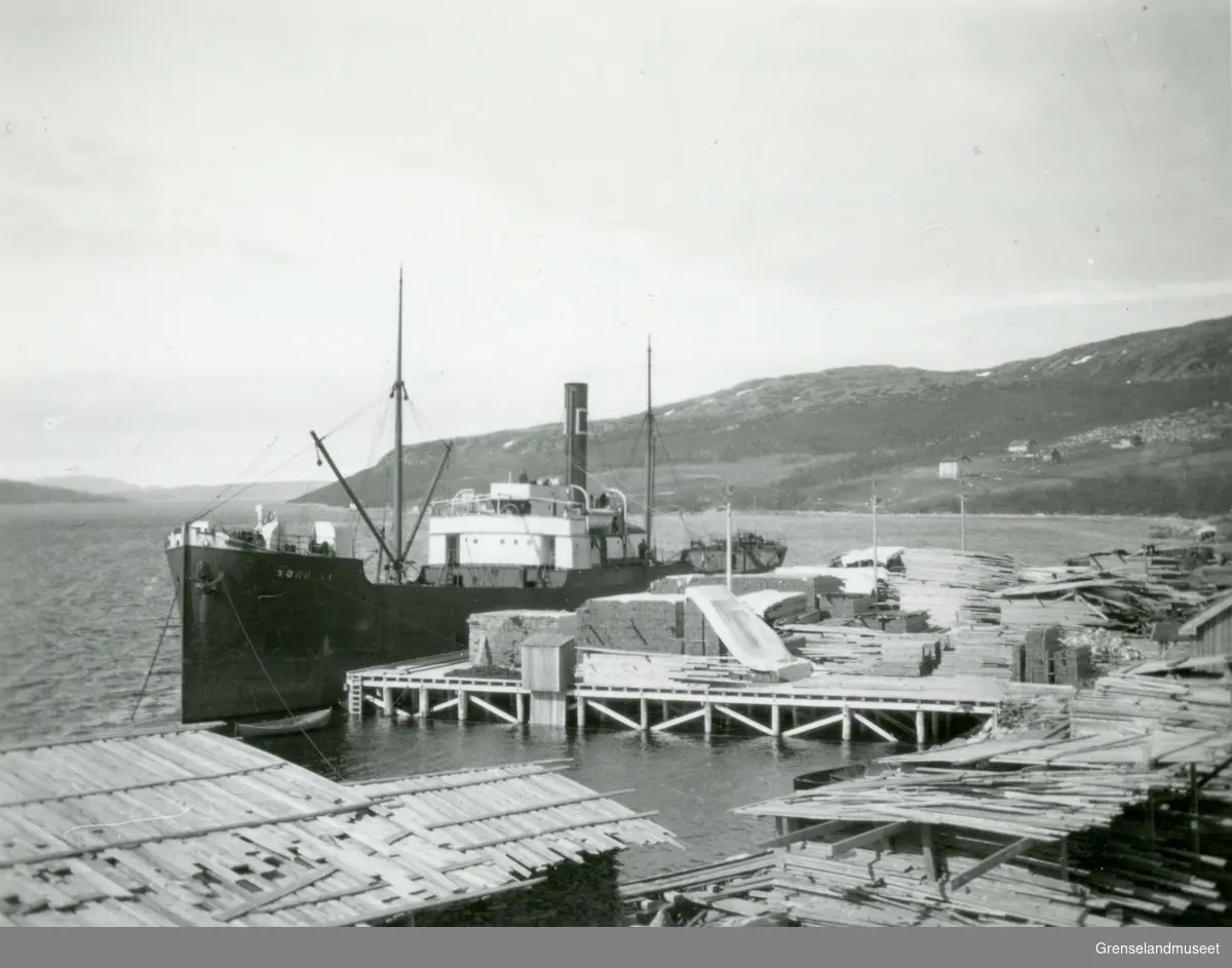 Pasvik Timber sitt anlegg på Jakobsnes før 1940. Lastekaien med skip og plankestabler i forgrunn. Utsikt over Bøkfjorden.
