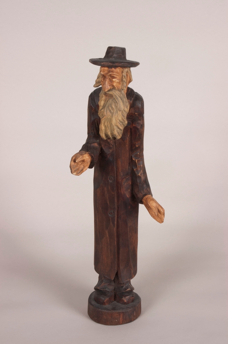 Eldre mann med utstrakt hånd. Har på seg en hatt og lang klesdrakt. Sko på bena. Mannen har halvlangt hår og langt skjegg til brystet.
