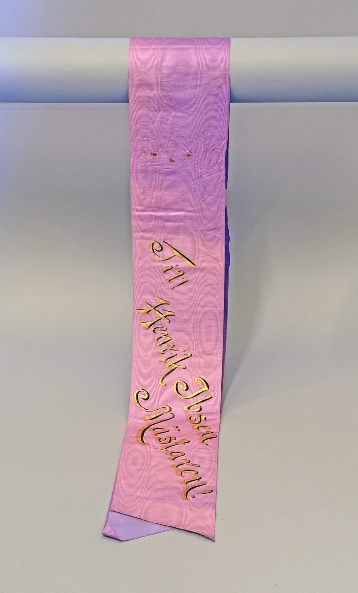 Båndet er lilla (rosa) med gulltekst - håndskrevet. 
