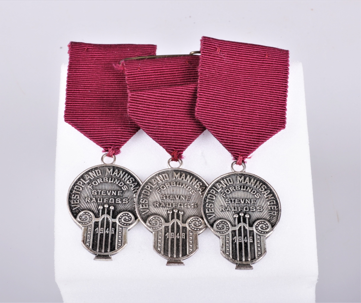Tre like minnemedaljer av sølvfarget metall med purpur festebånd. Innskift på medaljen