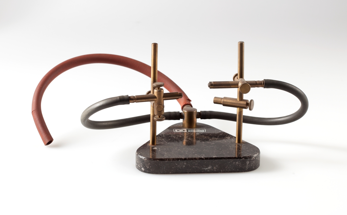 To gassbrennere montert mot hverandre på regulerbart stativ.
Slange for gass til hver av brennerne. Brennerne og gasstilkoblingen montert på trekantet marmorplate