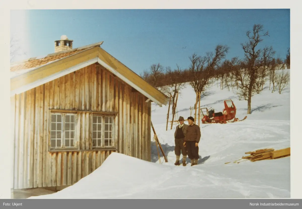 To menn med nikkers utendørs i snøen ved siden av husvegg. Bak sees en rød snøskuter