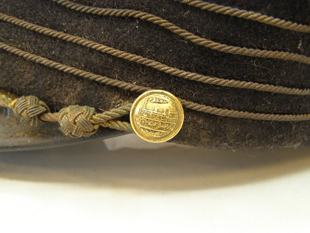 Skärmmössa  med kort skärm, med fyra guldfärgade snoddar och emaljerad A-loksknapp med monogram ovanför.
Guldfärgad stormträns och guldfärgade mösstränsknappar med 1899 års A-lok.
