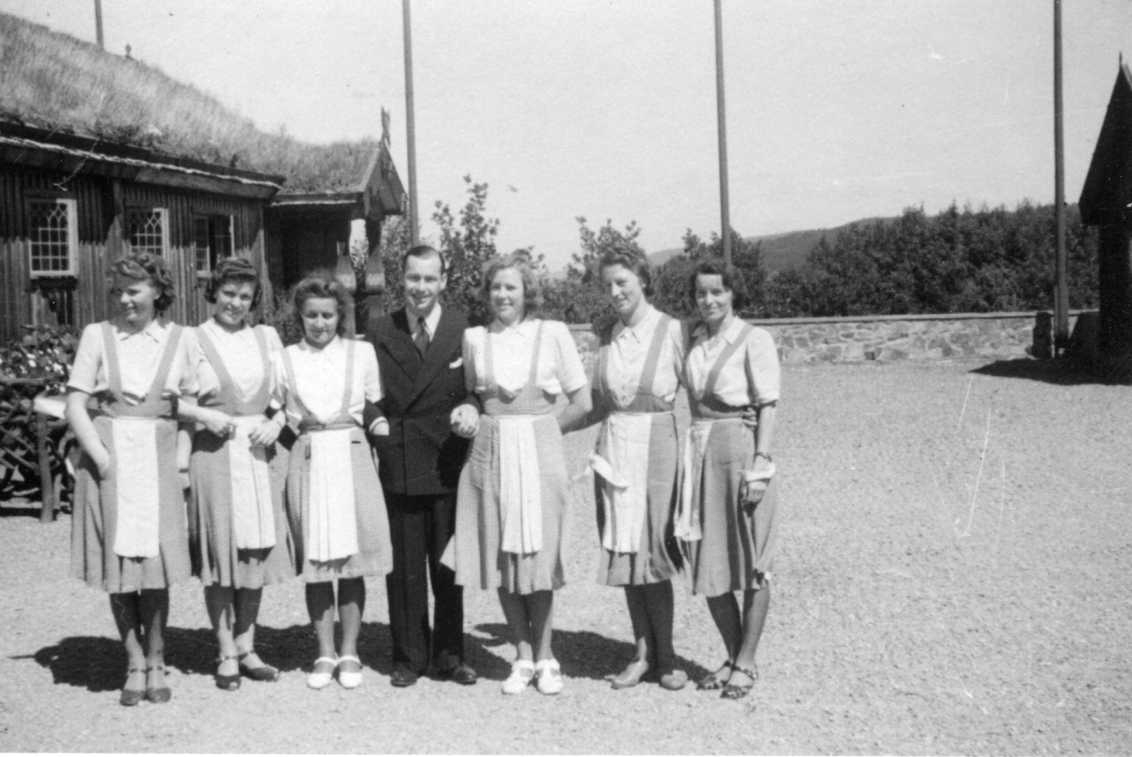 En man med dubbelknäppt kostym står på planen utanför Gyllene Uttern omgiven av tre kvinnor på vardera sidan. Han står armkrok med de närmaste. Kvinnorna är kortärmad ljus blus, kjol och förkläde. Det är sommar.