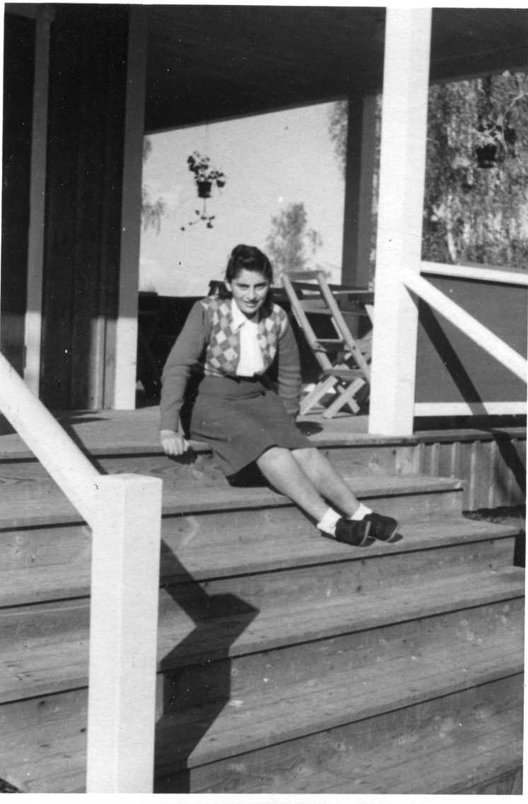 En ung polsk kvinna sitter på verandatrappa. Hon har en på bröstet rombmönstrad kofta med enfärgade armar ovanpå en ljus blus, kjol och ljusa strumpor i mörka skor.