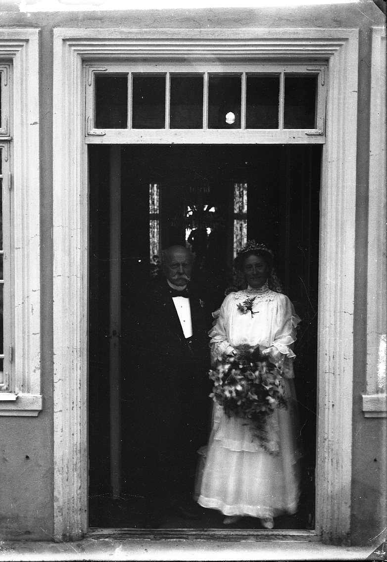 Bröllopsfotografi med Elsa Sofie von Otter och Adam Magnus Emanuel Lagerberg. Hon bär en ljus bröllopsklänning, ett diadem och slöja och håller en blombukett i händerna. Han bär frack. De står sannolikt vid entrén till Ribbagården.