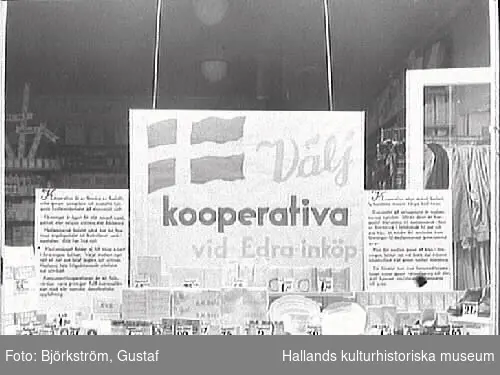 Skyltfönster till Kooperativas butik i Varberg. "Välj kooperativa vid Edra inköp" står det på en stor skylt i fönstret. I bakgrunden syns en stång med kläder.