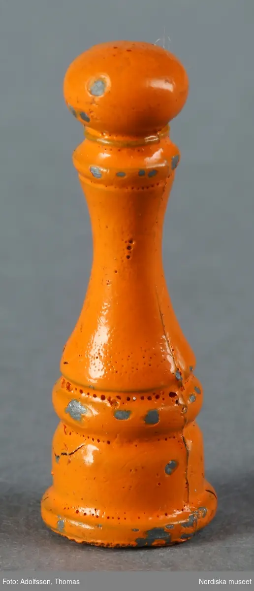 a) En vit plastburk med salt samt b) en orange pepparkvarn av metall. Hör till köksinredningen på första våningen i dockskåp NM.0331721+.
