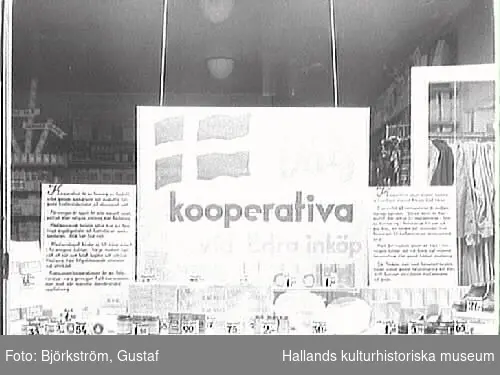 Skyltfönster till Kooperativa AB, butik i Varberg. "Välj kooperativa vid Edra inköp" står det på en stor skylt i fönstret. I bakgrunden syns en stång med kläder.