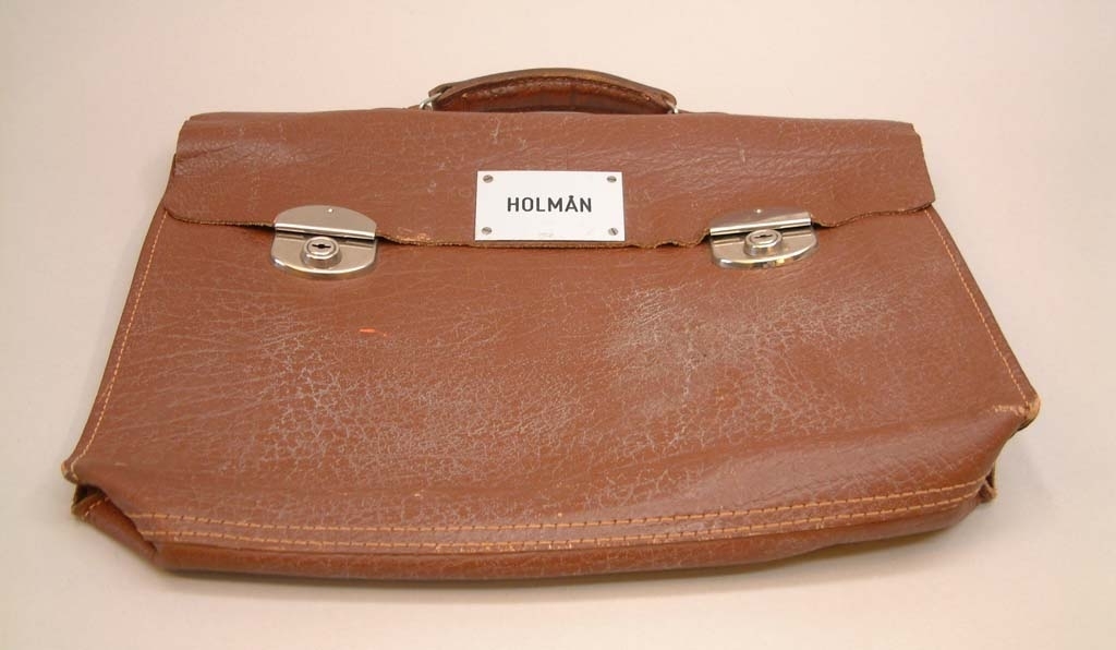 Brun portfölj i läder med två lås.
Två nycklar ligger i portföljen.
Metallskylt med text "HOLMÅN"