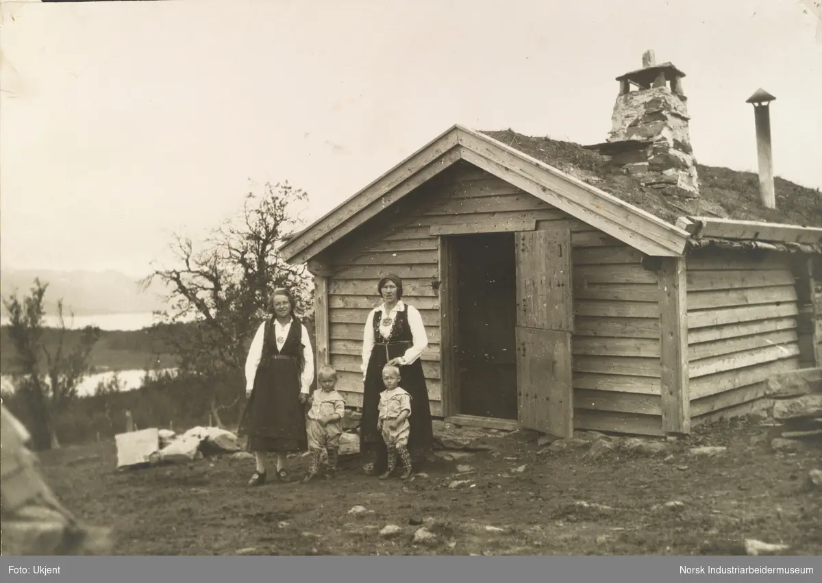 To kvinner i bunad med barn foran døråpning til hytte i fjellet