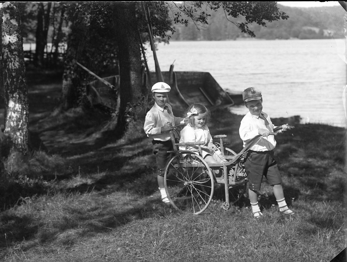 Barnen Stolpe från Stockholm leker vid en strand. Två pojkar kör en liten vagn i vilken en liten flicka sitter. Alla barn i fina kläder.