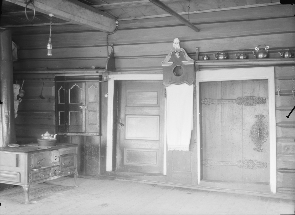 Interiør, kjøkken med tømmervegger, to utgangsdører med bostkast i mellom. Over ene døra står kaffekjeler. Innebygd skap til venstre med jernovn