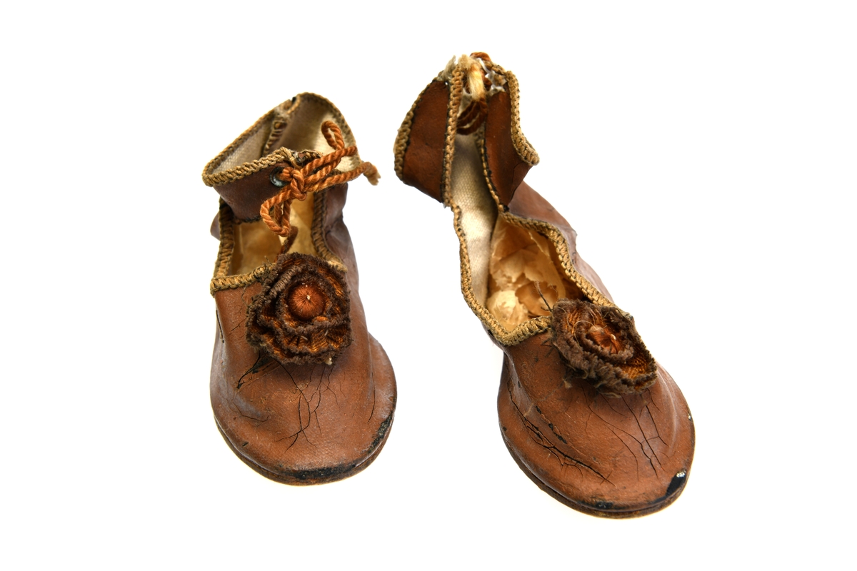 Et par dukkesko av tykk bomullslerret med lærsåle. Overdelen av skoen er av brunmalt bomullslerret. De er høy ved ankelen/bak og har ankelreimer som knytes med silkesnor foran. Kanten langs ankelreimene og åpningen på skoen er maskinsydd. Sømmen bak på skoene er håndsydd. Foran på skoene er det sydd på brune silkerosetter. Det er såle av papp i skoene. Det er limt på lærsåle på undersiden.