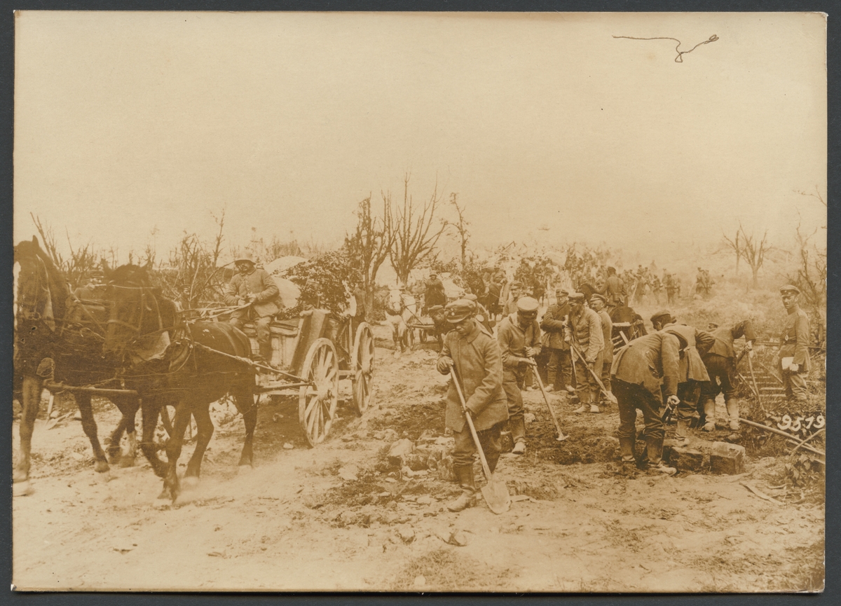 Bilden visar en trängkolonn med hästvagnar som kör på en landsväg. Bredvid dem arbetar en grupp soldater med skyfflar för att laga vägen.