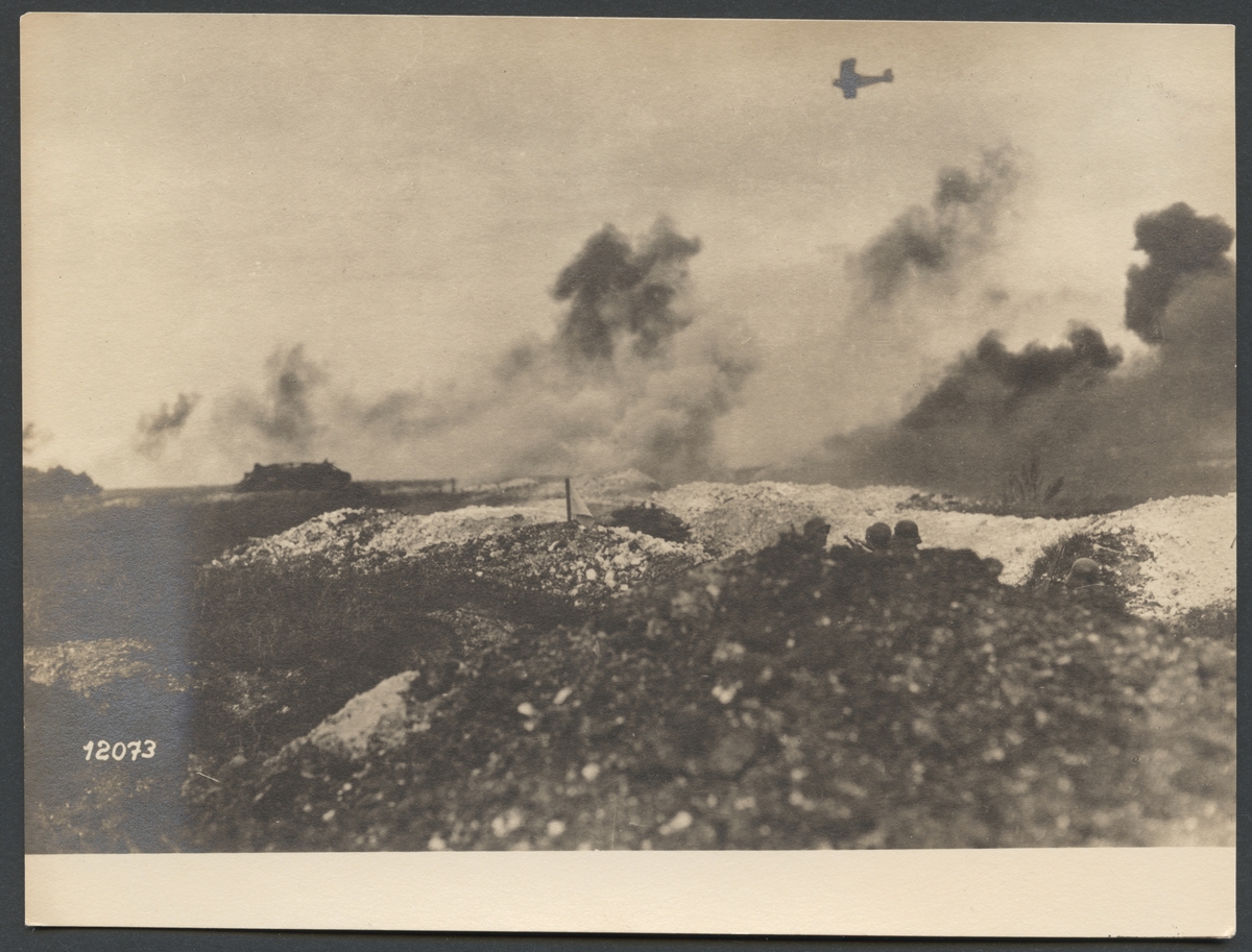 Bilden visar soldater som tar skydd i en skyttegrav och iakttar en stridsvagn som rör sig på slagfältet. Det syns flera explosioner från artillerield. Överst i bilden ser man ett spaningsflygplan.