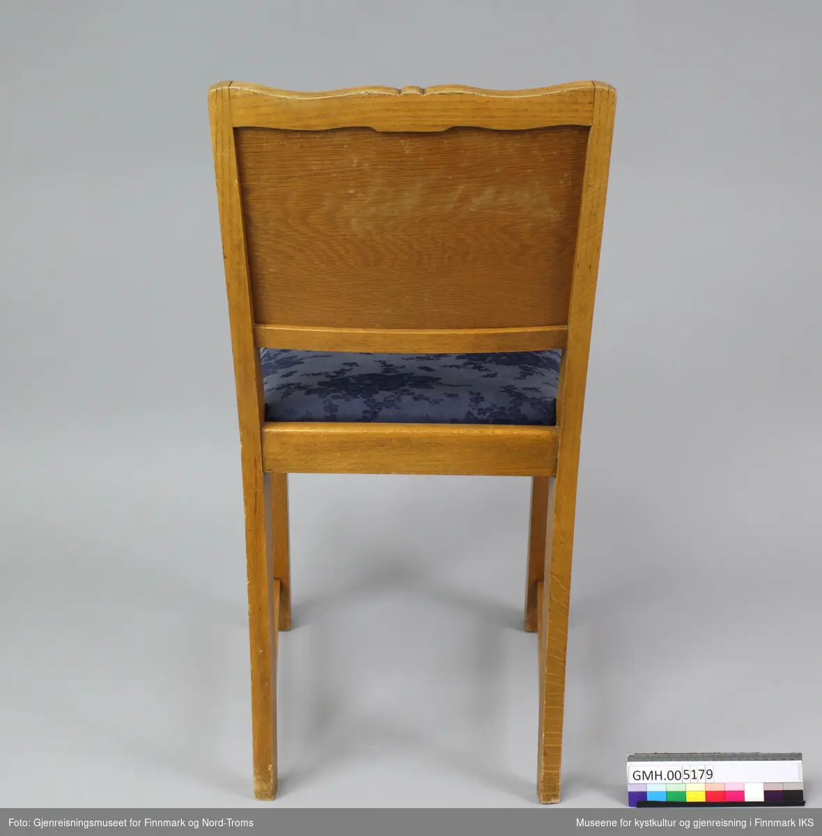 Spisebordstolen har en heldekkende rygg øverst med åpning nede ved setet. Ryggen er buet oppe med utskåret ornament. Rygglenet består av ramme med fyll med speilvendt finér. Skråmønstre i treverket vender ned mot midten av ryggen. Bakbena er sammenhengende fra  ryggstø til fot og buer innover mot setet. Forbena er firkantet.  Enkeltdelene stolramma består av er sammenføyd med tappe og hull-forbindelser. Stolen har trekantede klosser skrudd under setet ved bakbena. Stolsetene har polstring med et blå blomstermønstret tekstiltrekk . Trekket er stiftet fast under platen. Setet er løst og kan tas av.