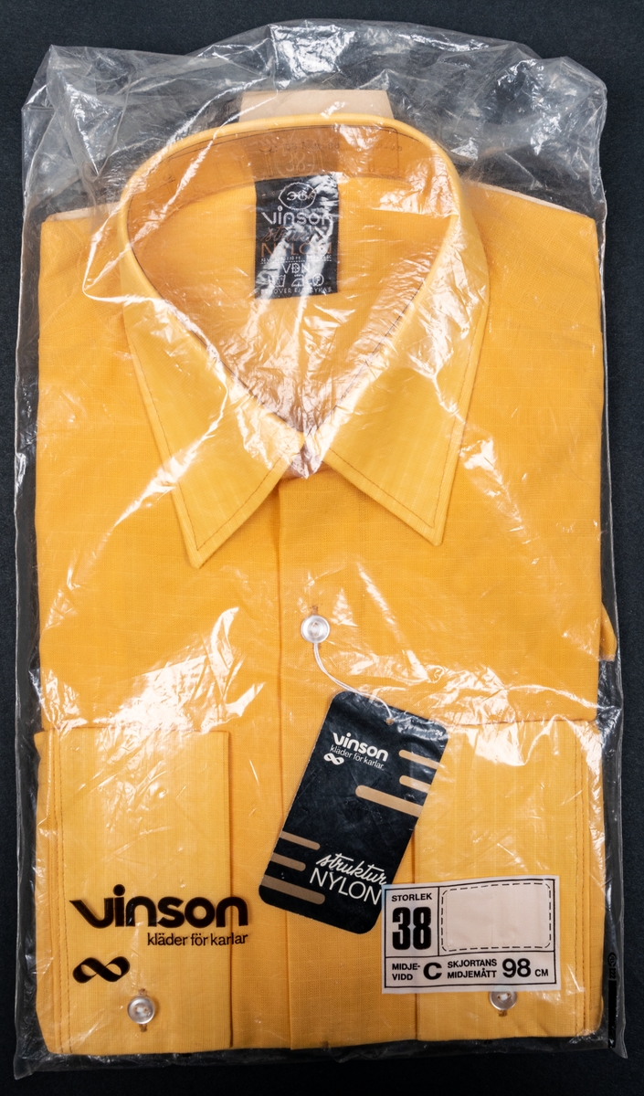 Gul nylonskjorta i originalförpackning av märket Vinson. Storlek 38. Ligger i plastpåse som också är i original. Svart märkeslapp i halsen med storlek och tvättanvisning. Måtten avser skjortan i vikt tillstånd.